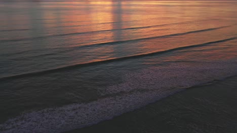 Luftdrohnenkamera-Bewegt-Sich-Bei-Sonnenaufgang-Am-Strand-Von-Unten-Nach-Oben,-Mit-Wellen,-Goldenem-Stundenlicht,-Gelb-Und-Rosa-Leuchtenden-Wolken,-Die-Sich-Im-Wasser-Spiegeln