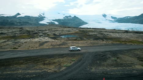 A-car-driving-on-a-famous-Icelandic-lave-fields-road-with-a-view-of-glaciers-and-snowcapped-mountains-in-the-background,-Iceland,-slow-motion-drone-shot