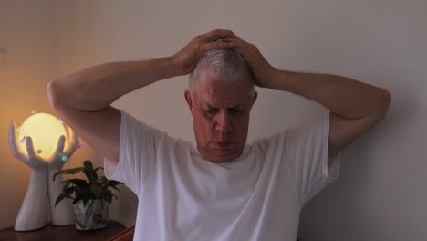 Exasperated-grey-haired-older-caucasian-man-in-white-T-shirt-holds-head-in-frustration