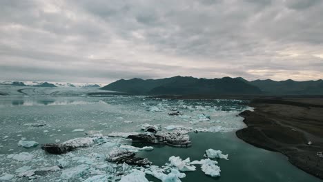 Scenic-view-of-iceberg-drifting-on-the-glacier-water-surface,-Melting-glacier-due-to-global-warming,-Mountains-under-an-overcast-sky-in-the-background,-Iceland,-Slow-motion-drone-shot
