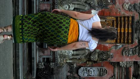 Tiro-Vertical-En-Cámara-Lenta-De-Una-Mujer-Joven-Vestida-Con-Un-Pareo-Verde-Caminando-Hacia-La-Entrada-De-Un-Templo-En-Bali,-Indonesia