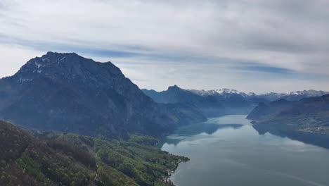 Pristine-Wilderness-of-Alps,-Aerial-View-of-Mountain-Hills-and-Peaks-Above-Picturesque-Lake-on-Spring-Season