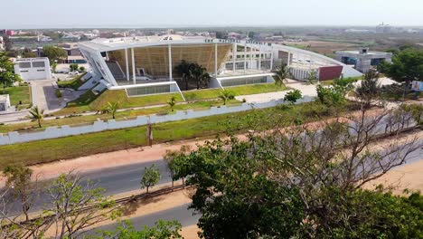 Absteigende-Drohnenaufnahme-Des-Gebäudes-Der-Nationalversammlung-Von-Gambia-Mit-Blick-Auf-Die-Stark-Befahrene-Straße-Mit-Bäumen-Im-Vordergrund-An-Einem-Sonnigen-Sommertag