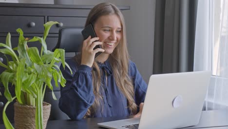Feliz-Mujer-Emocionada-Que-Trabaja-Desde-La-Oficina-Habla-Por-Teléfono-Mientras-Usa-Una-Computadora-Portátil