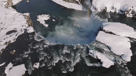 Waterfall-in-Winter-Landscape-of-Iceland,-Revealing-Drone-Shot,-Flowing-Water,-Snow-and-Rainbow-Above-Canyon
