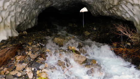 Aerial-view-entering-dark-glacial-ice-cave-over-cascading-stream-water-rushing-from-tunnel-passage-in-Provo,-Utah