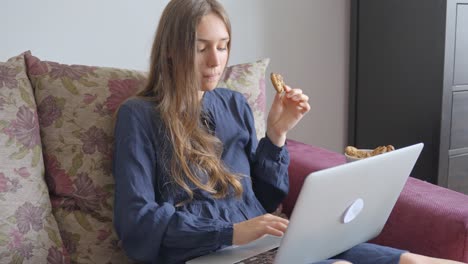 Young-woman-working-from-home,-sitting-on-the-sofa-with-a-laptop-and-snacking-on-cookies,-in-Slow-Motion