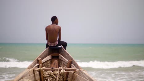 Rückansicht-Eines-Nicht-Identifizierbaren-Afrikanischen-Mannes,-Der-Entspannt-Am-Rand-Eines-Kanus-Sitzt,-Auf-Das-Meer-Blickt-Und-Seine-Hände-öffnet