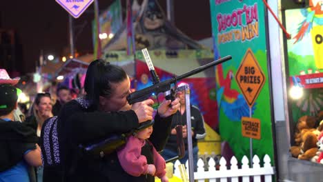 Mom-with-a-baby-in-the-front-playing-carnival-shooting-gallery-game-with-recreational-guns-loaded-with-silicon-rubber-balls-at-iconic-Ekka,-Royal-Queensland-Show-at-Brisbane-Showgrounds
