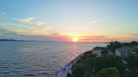 Lift-up-drone-footage-over-tree-canopy-and-red-rooftops-in-sunset-over-old-town-Zadar,-Croatia-with-promenade-and-large-amount-of-tourists-walking