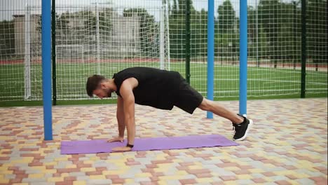 Hombre-En-Ropa-Deportiva-Negra-Haciendo-Flexiones-En-La-Alfombra-Con-Campo-De-Fútbol-En-El-Fondo