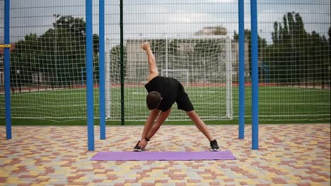 Hombre-Con-Ropa-Deportiva-Negra-Haciendo-Ejercicios-De-Calentamiento-En-Una-Alfombra-Con-Un-Campo-De-Fútbol-En-El-Fondo,-Ejercicio-De-Molino