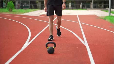 Close-up-footage-of-a-man-in-black-sportswear-comes-to-train-with-weight-bob-outdoors-on-the-racetrack-outdoors