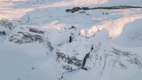 Aerial-panoramic-view-of-a-winter-landscape-covered-in-snow,-over-Skogafoss-waterfall,-in-Iceland,-at-sunset