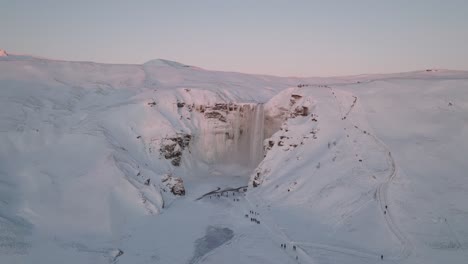 Aerial-panoramic-view-of-a-winter-landscape-covered-in-snow,-of-Skogafoss-waterfall,-in-Iceland,-at-sunset