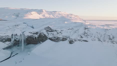 Aerial-panoramic-view-of-a-winter-landscape-covered-in-snow,-over-Seljalandsfoss-waterfall,-in-Iceland,-at-dusk