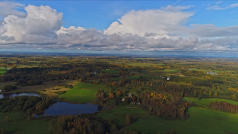 Timelapse-aerial-majestic-view-of-a-countryside-of-fields,-trees-and-a-lake-as-low-lying-clouds-roll-through-the-blue-sky