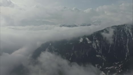 Aerial-view-of-partially-snow-covered,-tree-filled-mountain-tops-above-thick-cloud-cover