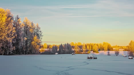 Timelapse-of-farmland,-trees-and-hay-bales-covered-with-snow,-as-the-sun-reflects-off-the-landscape-and-the-trees-gently-blow-in-the-breeze