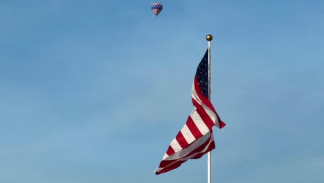 Low-angle-portrait-of-American-flag-swaying-against-blue-sky,-distant-hot-air-balloon,-closeup