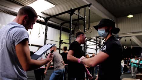 Tres-Jóvenes-Caucásicos-Musculosos-Golpean-Los-Puños-Para-Saludarse-Durante-Una-Competencia-De-Crossfit-En-Un-Gimnasio-Con-Otras-Personas-Detrás-De-Ellos-Y-Equipo-De-Pesas