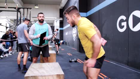 Un-Joven-Caucásico-Realizando-Cajas-De-Salto-En-Un-Gimnasio-De-Crossfit-Bajo-La-Supervisión-De-Un-Evaluador-Durante-Una-Competición-De-Crossfit