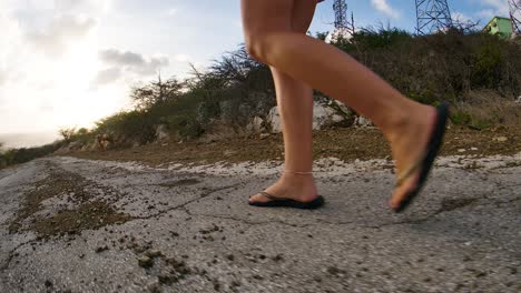 Young-Caucasian-female-feet-tracking-shot-low-to-ground-over-eroded-old-broken-asphalt-road,-downhill-sunset-in-backrgound