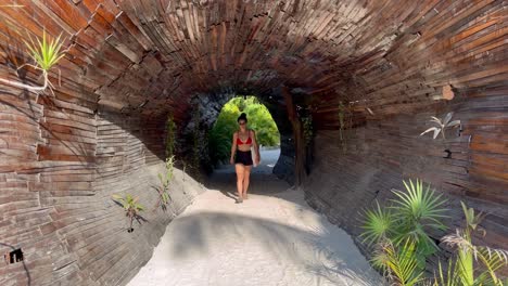 A-young-woman-in-a-bikini-walking-through-a-wooden-tunnel-on-Tulum-Beach-in-Zona-Hotelera,-Selina-Hotel