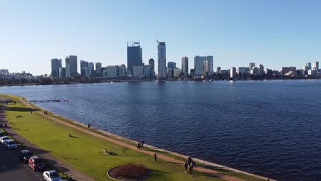 Drone-shot-rising-over-and-South-Perth-foreshore-Swan-River-to-skyline-of-Perth,-Western-Australia-and-lawn