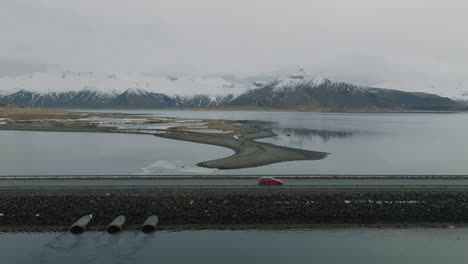 Aerial-View-of-Red-Car-Moving-on-Road-on-Embankment-Above-Glacial-Lake-With-Snow-Capped-Volcanic-Hills-in-Background,-Iceland