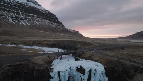 Drone-Shot-of-Man-Walking-on-Bridge-Above-Glacial-River-and-Waterfall-in-Landscape-of-Iceland-On-Cold-Spring-Day