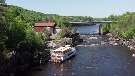Descending-close-up-aerial-shot-of-a-riverboat-docked-on-the-Saint-Croix-River-in-Taylors-Falls,-Minnesota