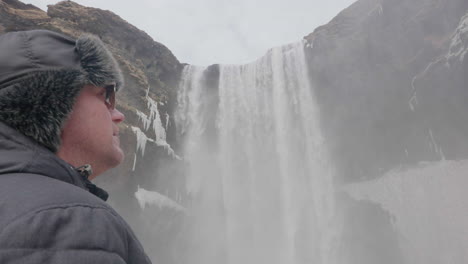 Middle-Age-Man-in-Warm-Winter-Clothes-Standing-Under-Skógafoss-Waterfall,-Natural-Landmark-of-Iceland,-Close-Up