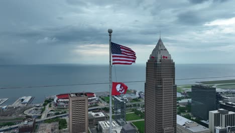 Cleveland,-Ohio,-Estados-Unidos,-Vista-Aérea-De-La-Bandera-Nacional-Estadounidense-Ondeando-En-El-Poste-Frente-A-La-Torre-Clave-Y-La-Costa-Del-Lago-Erie
