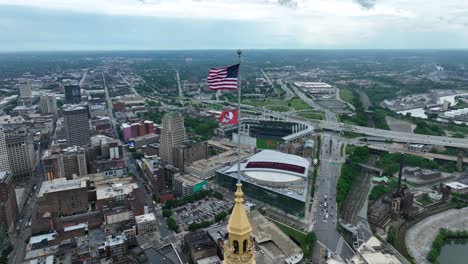 Cleveland,-Ohio,-USA,-Hochhaus-Drohnenaufnahme-Einer-Amerikanischen-Flagge-Und-Einer-Guardians-Flagge,-Die-An-Einer-Stange-Weht,-Mit-Der-Rocket-Mortgage-Arena-Und-Dem-Progressive-Field-Baseballstadion-Im-Hintergrund