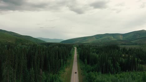 Drone-aerial-tilt-shot-of-a-lone-vehicle-speeding-along-a-highway-amidst-a-green-forest-in-Alaska