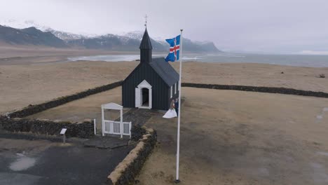 Vista-Aérea-De-Una-Pareja-De-Novios-Frente-A-La-Iglesia-Negra-En-Búðir,-Islandia,-Toma-De-Drones-De-Un-Punto-De-Referencia-Y-Bandera-Nacional,-50-Fps