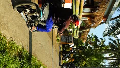Vertical-slow-Motion-shot-of-Balinese-construction-worker-driving-to-construction-site-with-bamboo-canes-on-his-shoulder-while-riding-his-scooter