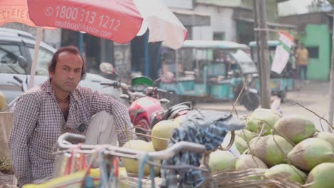 Asian-Coconut-seller-selling-coconuts-in-footpath-stall-on-roadside-at-daytime,-looking-at-camera