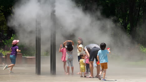 Un-Grupo-De-Niños-Se-Divierte-Refrescándose-En-Un-Caluroso-Día-De-Verano-Bajo-Un-Sistema-De-Enfriamiento-Por-Goteo-De-Microflujo-En-Un-Parque-Público,-Rociando-Niebla-Aero-Fría---Bosque-Ciudadano-De-Yangjae