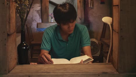 Looking-through-an-open-window-at-a-young-Asian-male-reads-a-book-at-a-desk-in-a-rustic-room