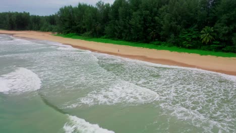 Aerial-drone-camera-close-up-showing-huge-waves-coming-from-the-sea-to-the-beach-with-a-forest-in-front-and-a-man-walking-alone-on-a-seedling