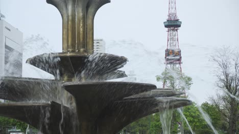 Soplando-Agua-En-La-Fuente-Del-Parque-Odori-Con-La-Torre-De-Televisión-De-Sapporo-Al-Fondo-En-Hokkaido,-Japón