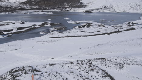 Slow-aerial-revealing-shot-of-a-half-frozen-lake-at-the-base-of-Icelandic-mountains
