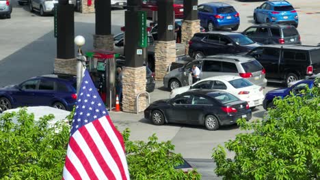 Die-Amerikanische-Flagge-Weht-Im-Wind,-Während-Während-Der-Gaskrise-Menschen-In-Ihren-Autos-Schlange-Stehen