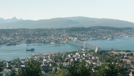 Stunning-Aerial-View-of-The-City-of-Tromsø-on-a-Sunny-Summer-Day,-with-some-Traffic-on-the-Bridge-and-a-Cargo-Ship-in-the-Foreground,-Norway