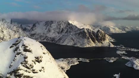 Aerial-view-backwards-over-the-snowy-Reinebringen-peak,-with-the-Reine-town-in-the-background,-in-Norway
