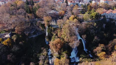 Drone-shot-of-beautiful-Edessa-Waterfall-in-forest-landscape-during-Sunny-day-and-town-in-background,-Greece