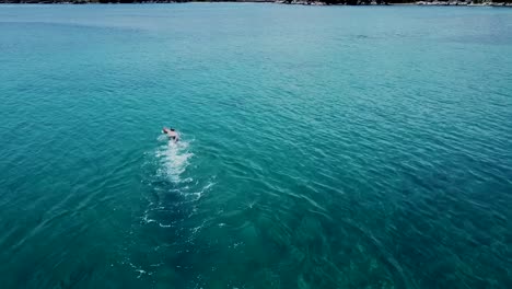 Aerial-tilt-up-shot-of-swimmer-in-clear-water-in-bay-of-Greece-with-mountains-in-background-during-sunny-day