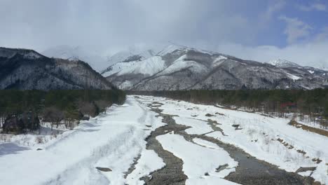 gliding-forward,-the-drone-captures-the-beauty-of-a-winter-river-in-Hakuba-valley,-Japan,-the-icy-flow-carves-its-way-through-the-snowy-banks-in-a-beautiful-mountains-landscape